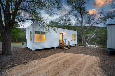 Modern white tiny cabin on stilts with wooden steps and warm interior glow, parked on a cleared dirt pad among oak trees on a wooded hillside at twilight