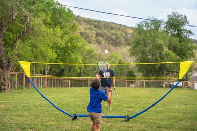 Young boy playing badminton with an adult across a portable yellow net on a grassy outdoor lawn, trees and rolling hills in the background — family-friendly park scene.