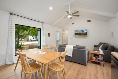 Bright open-concept living and dining area with light wood table and chairs, gray sofas, vaulted ceiling and fan, wall-mounted TV, and a large sliding door framing oak trees and a yard — modern minimalist home interior.