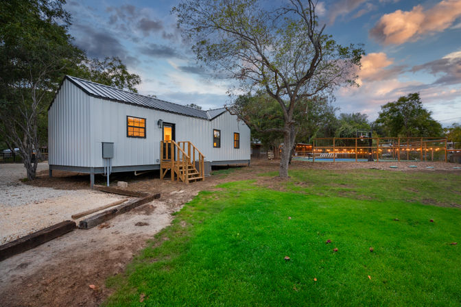 Dusk scene of a white metal-clad cabin on stilts with wooden steps and glowing windows, set on a green lawn with mature trees and a fenced, string-lit pool area with a basketball hoop.