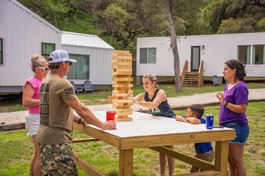 Group of adults and a child playing a tall giant Jenga tower on a raised wooden table outside white cabin units in a grassy campground on a sunny day.