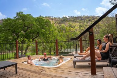 Group of friends relaxing in an outdoor hot tub on a wooden deck with trees and a scenic green hillside under a sunny blue sky