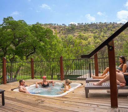 Group of friends relaxing in an outdoor hot tub on a wooden deck with trees and a scenic green hillside under a sunny blue sky