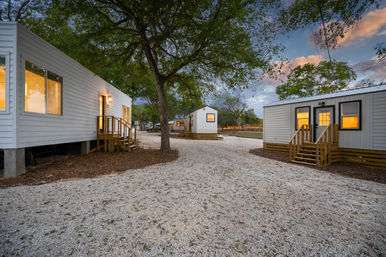 Cozy tiny cabin rentals arranged around a gravel courtyard under large oak trees at dusk, warm interior lights glowing.