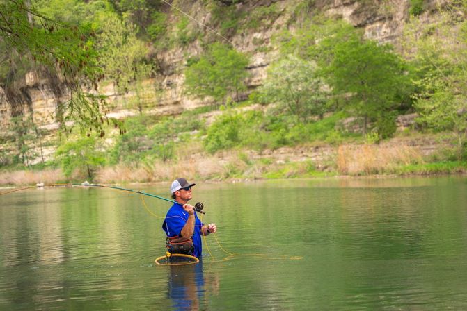 Angler fly-fishing waist-deep in a green river, casting a line near limestone bluffs and spring foliage