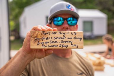 Person wearing a cap and mirrored sunglasses holds a wooden block outdoors with a handwritten joke about gravity: 'There's one thing in life that's a constant and always brings you down... Gravity.' Blurred small buildings and people in the background.