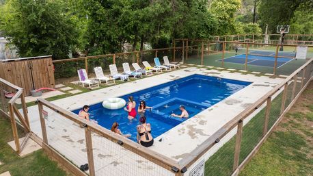 Fenced rectangular outdoor pool with a small group chatting in the water, inflatable ring, row of lounge chairs on the concrete deck, and an adjacent fenced multi-sport court and basketball hoop surrounded by trees.