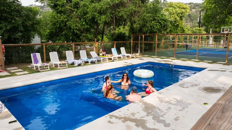 Group of six people chatting in a blue rectangular outdoor pool with a built-in bench and striped inflatable tube, concrete deck lined with white lounge chairs and towels, fenced yard with a tennis/basketball court and green trees and hills beyond.