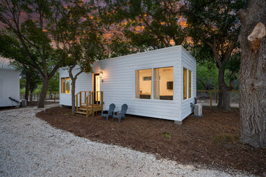 White modern tiny house cabin with warm interior lights, front steps and two outdoor chairs tucked among oak trees at a wooded glamping site at sunset