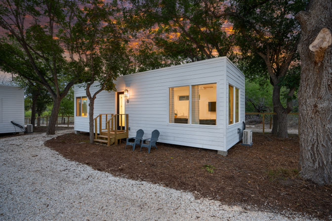 White modern tiny house cabin with warm interior lights, front steps and two outdoor chairs tucked among oak trees at a wooded glamping site at sunset