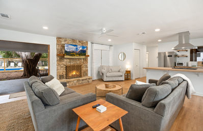 Cozy open-concept living room with stone fireplace and wall-mounted TV, two gray sofas around a round coffee table, hardwood floors and breakfast bar kitchen, sliding door to backyard with large tree and pool