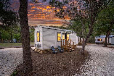 Cozy tiny white cabin with glowing windows and front steps on a tree-lined gravel lot at sunset, two Adirondack chairs outside and neighboring cabins along the path.