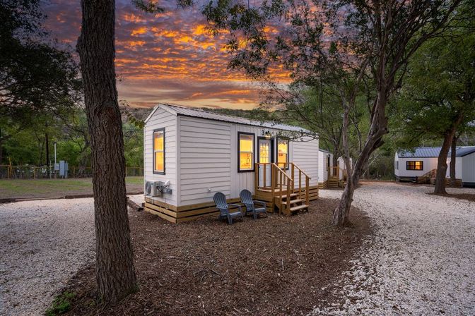 Cozy tiny white cabin with glowing windows and front steps on a tree-lined gravel lot at sunset, two Adirondack chairs outside and neighboring cabins along the path.
