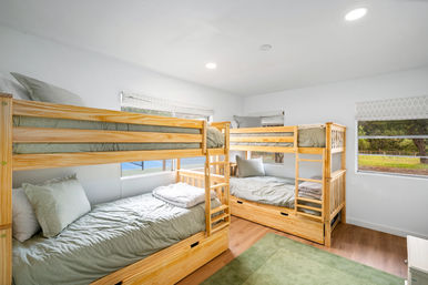 Sunlit bedroom with two natural-wood twin-over-twin bunk beds, soft sage green bedding and pillows, hardwood floors, a green area rug and windows overlooking trees.