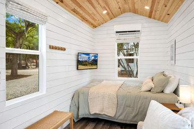 Cozy tiny house bedroom interior with white shiplap walls and wood-plank vaulted ceiling, sage linen bed with knit throw, wall-mounted TV, windows overlooking trees and a wicker bench.