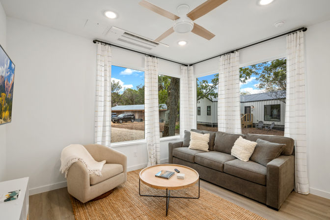 Bright, cozy modern living room with gray sofa, beige armchair, round wood coffee table and ceiling fan; large windows with white curtains overlook trees, a parked car and cabin-style buildings in a rural setting.