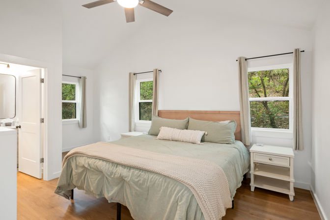 Cozy bright minimalist bedroom with king-size bed in sage-green bedding and knit throw, wooden headboard, two windows framing leafy forest views, white walls, ceiling fan and hardwood floors.