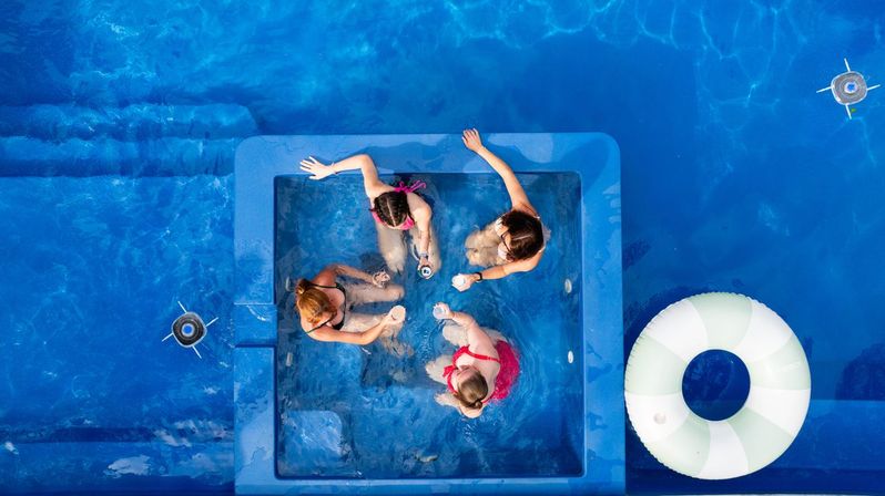 Aerial view of four people relaxing in a square spa pocket inside a bright blue swimming pool, holding drinks and chatting next to a white-and-green inflatable ring — sunny summer pool scene.