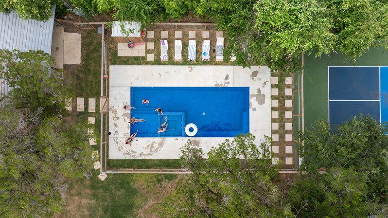 Aerial drone view of a rectangular backyard swimming pool with people lounging and swimming on a white deck, striped lounge chairs, surrounding trees and an adjacent blue tennis court