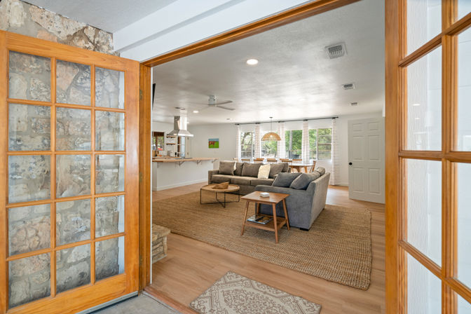 Sunlit open-concept living room and kitchen seen through wooden French doors, with a gray sectional on a woven jute rug, round coffee table and wooden side table, bar-style island with stools, large windows showing trees, hardwood floors and natural stone entry wall — bright, cozy neutral interior.