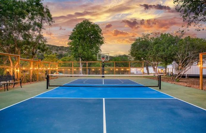 Blue outdoor tennis/pickleball court with center net and basketball hoop, wooden fence strung with lights, surrounded by trees and hills under a colorful sunset sky
