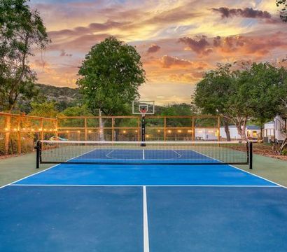 Blue outdoor tennis/pickleball court with center net and basketball hoop, wooden fence strung with lights, surrounded by trees and hills under a colorful sunset sky