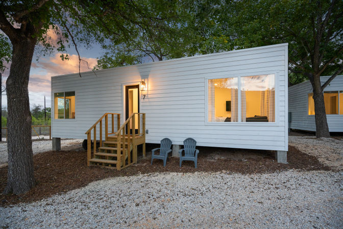 Dusk view of a modern white tiny cabin on stilts with wooden steps, two Adirondack chairs on gravel, glowing large windows, and surrounding trees.