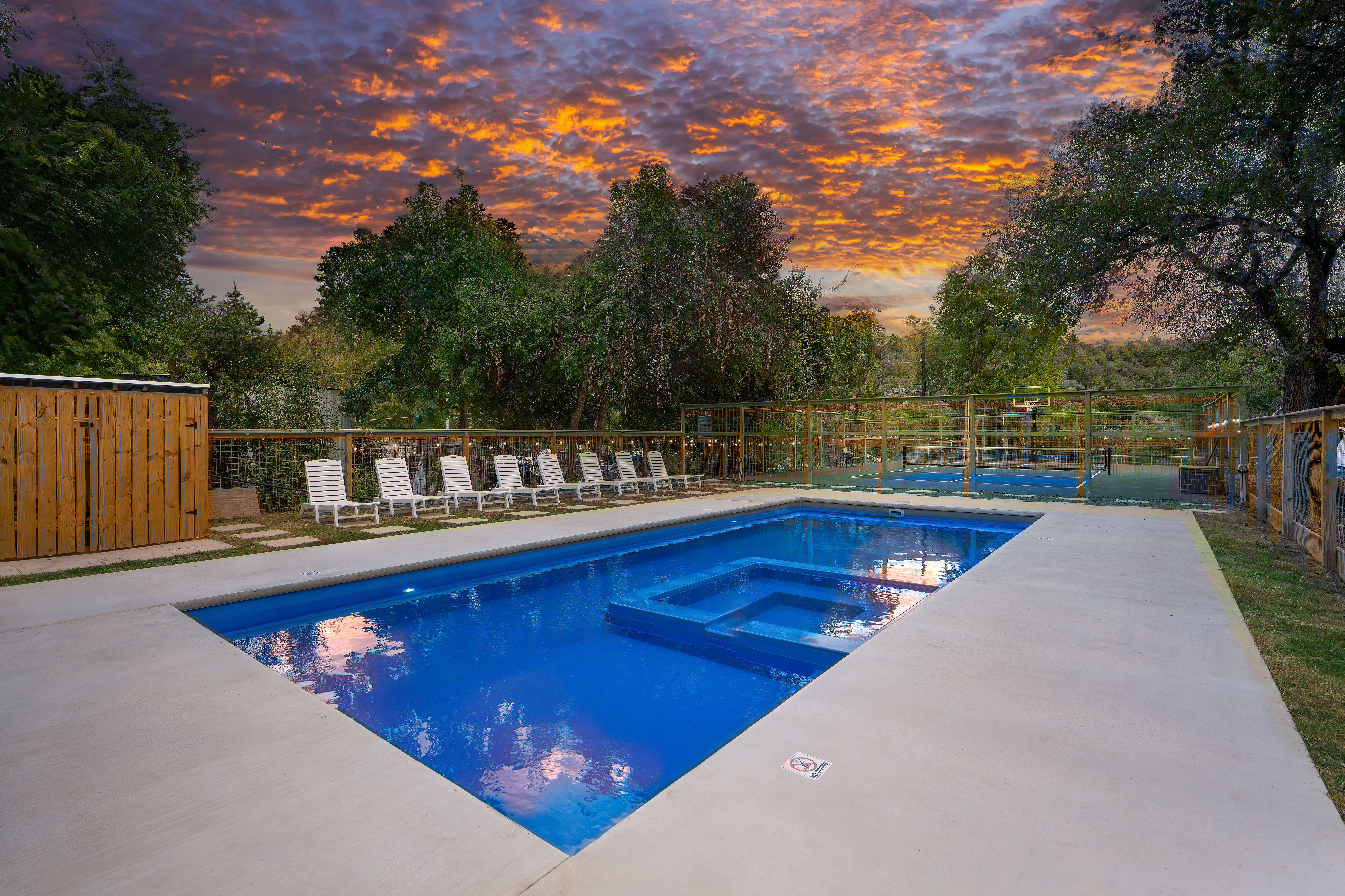 Blue rectangular outdoor pool with built-in spa on a concrete deck, row of white lounge chairs, fenced multi-sport court and trees under a dramatic orange sunset sky.