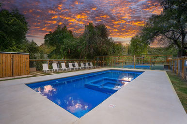 Blue rectangular outdoor pool with built-in spa on a concrete deck, row of white lounge chairs, fenced multi-sport court and trees under a dramatic orange sunset sky.