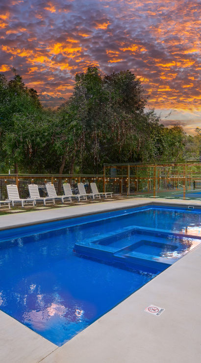 Blue rectangular outdoor pool with built-in spa on a concrete deck, row of white lounge chairs, fenced multi-sport court and trees under a dramatic orange sunset sky.