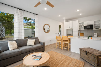 Bright modern open-plan living room and kitchen with a gray sofa, round wooden coffee table, white island, wooden dining chairs, woven rug and large window overlooking trees