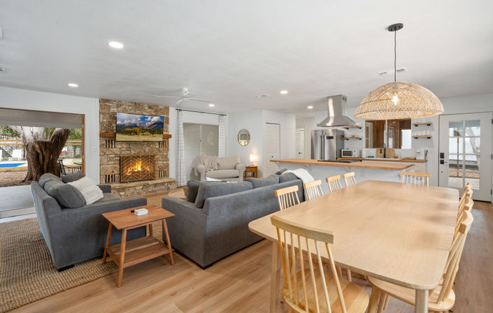 Open-concept living room and kitchen with light wood floors, gray sofas facing a stone fireplace and wall-mounted TV, large wooden dining table under a woven pendant light, breakfast bar with stools and stainless steel appliances, sliding doors opening to a yard with a large tree.