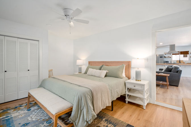 Bright, airy coastal-style bedroom with wooden headboard, sage-green bedding, knit throw and bench at foot, white nightstands with lamps, ceiling fan, louvered closet doors, hardwood floors and view into open-plan living room and kitchen.