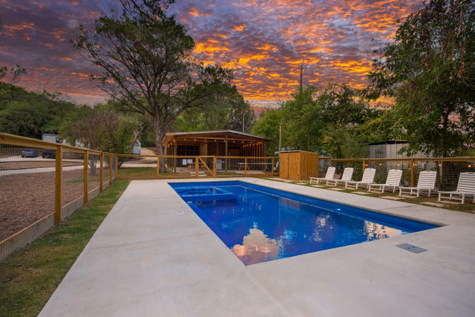 Rectangular outdoor swimming pool reflecting a fiery orange sunset, flanked by white lounge chairs, wooden fence and a rustic cabin pavilion among trees for a relaxed rural retreat.