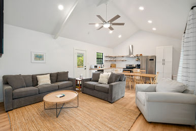 Bright open-plan living room and kitchen with vaulted white ceiling and ceiling fan, gray sofas and striped armchair around a round wooden coffee table on a jute rug, wooden dining table, and stainless-steel appliances with open shelving.