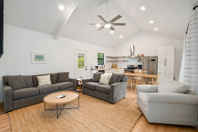 Bright open-plan living room and kitchen with vaulted white ceiling and ceiling fan, gray sofas and striped armchair around a round wooden coffee table on a jute rug, wooden dining table, and stainless-steel appliances with open shelving.