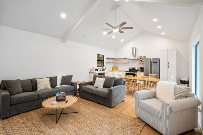 Cozy, bright open-plan living room and kitchen with vaulted ceiling and ceiling fan; gray sofas and striped armchair around a round wooden coffee table on a jute rug, wooden dining table with chairs, white cabinets and stainless-steel appliances.