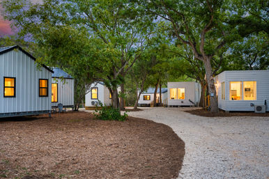 Dusk view of modern tiny-home cabins in a wooded retreat — warm-lit windows, gravel pathway winding between trees and mulched grounds, perfect for a glamping nature getaway.