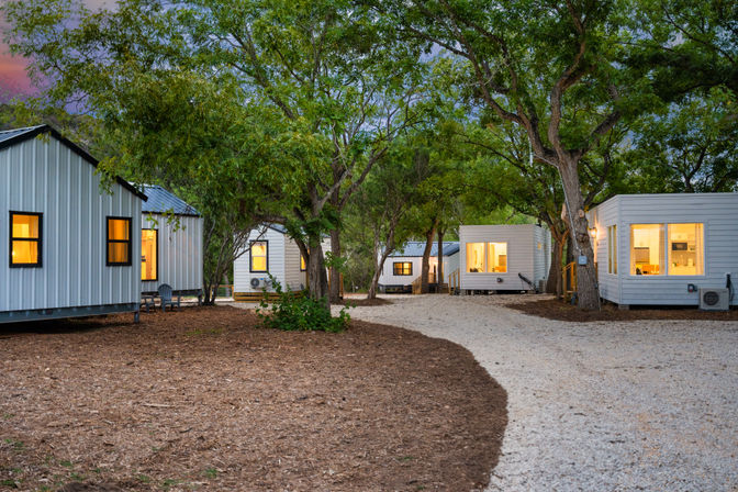 Dusk view of modern tiny-home cabins in a wooded retreat — warm-lit windows, gravel pathway winding between trees and mulched grounds, perfect for a glamping nature getaway.