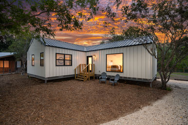 Sunset-lit modern metal-clad tiny house on a wooded lot, L-shaped raised layout with wooden steps, two outdoor lounge chairs on mulch, and a large glowing window revealing a cozy bedroom beneath a dramatic orange-pink sky.