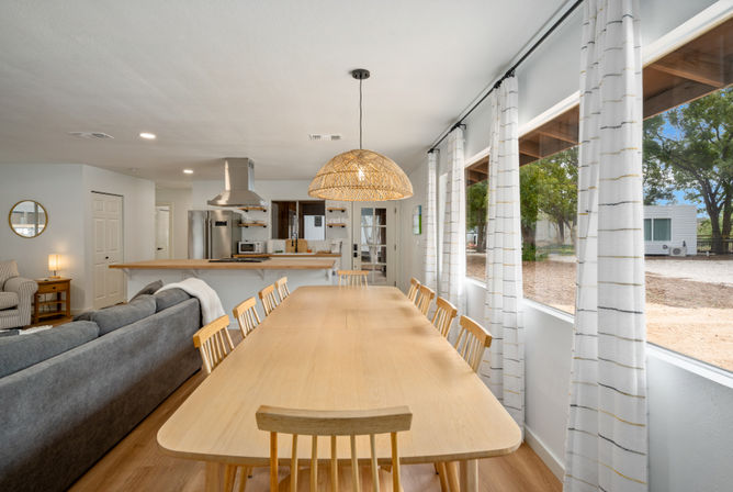 Bright open-plan dining and kitchen in a modern farmhouse-style home: long light-wood table with matching chairs under a woven rattan pendant, gray sofa by the kitchen island with stainless appliances, and large windows with white curtains overlooking a tree-lined yard.