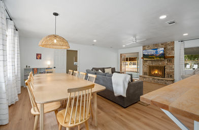 Sunlit open-plan living and dining room with a long light-wood table and chairs under a woven pendant, gray sectional sofa, stone fireplace with wall-mounted TV, hardwood floors and sliding doors to a pool