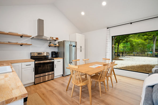 Bright open-concept kitchen and dining area with light wood counters and flooring, stainless steel range and fridge, floating shelves, six-seat wooden dining table, and large picture window overlooking a leafy fenced backyard.