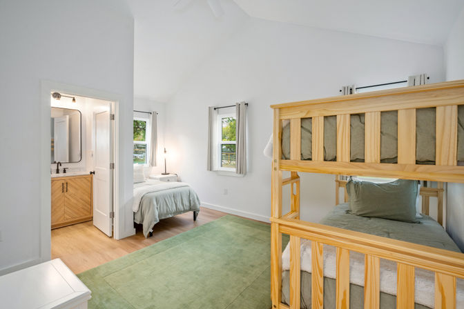 Cozy bright bedroom with natural-wood bunk beds, single bed, white walls, hardwood floors and green area rug, soft natural light from two windows and an en-suite bathroom visible through an open door.