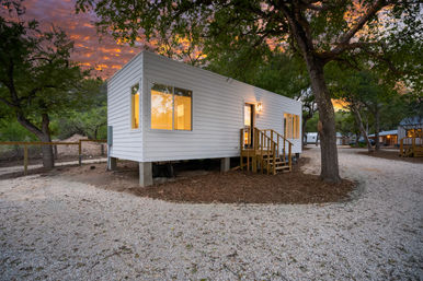 Modern white tiny house on concrete piers with wooden steps and warm glowing windows, tucked among oak trees at a wooded campground with a gravel path under a colorful sunset sky