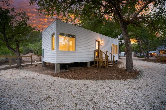 Modern white tiny house on concrete piers with wooden steps and warm glowing windows, tucked among oak trees at a wooded campground with a gravel path under a colorful sunset sky