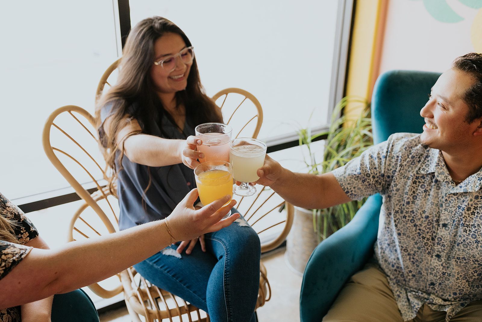 Three friends toasting colorful drinks in a bright, cozy cafe with rattan chairs and houseplants