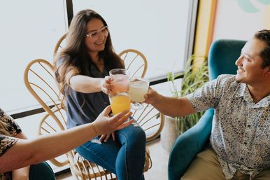 Three friends toasting colorful drinks in a bright, cozy cafe with rattan chairs and houseplants