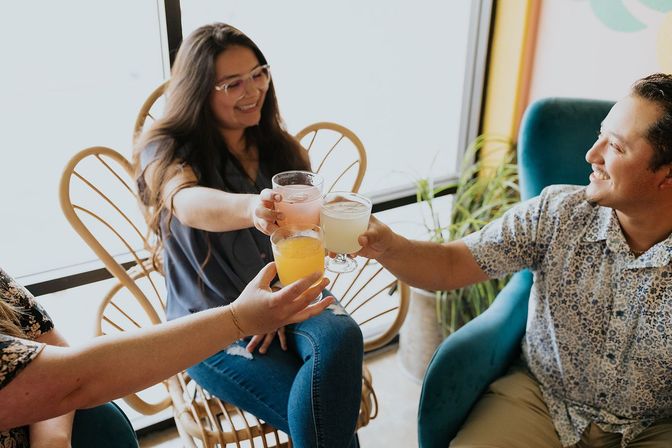 Three friends toasting colorful drinks in a bright, cozy cafe with rattan chairs and houseplants