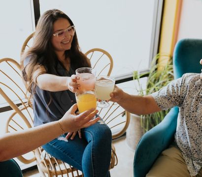 Three friends toasting colorful drinks in a bright, cozy cafe with rattan chairs and houseplants